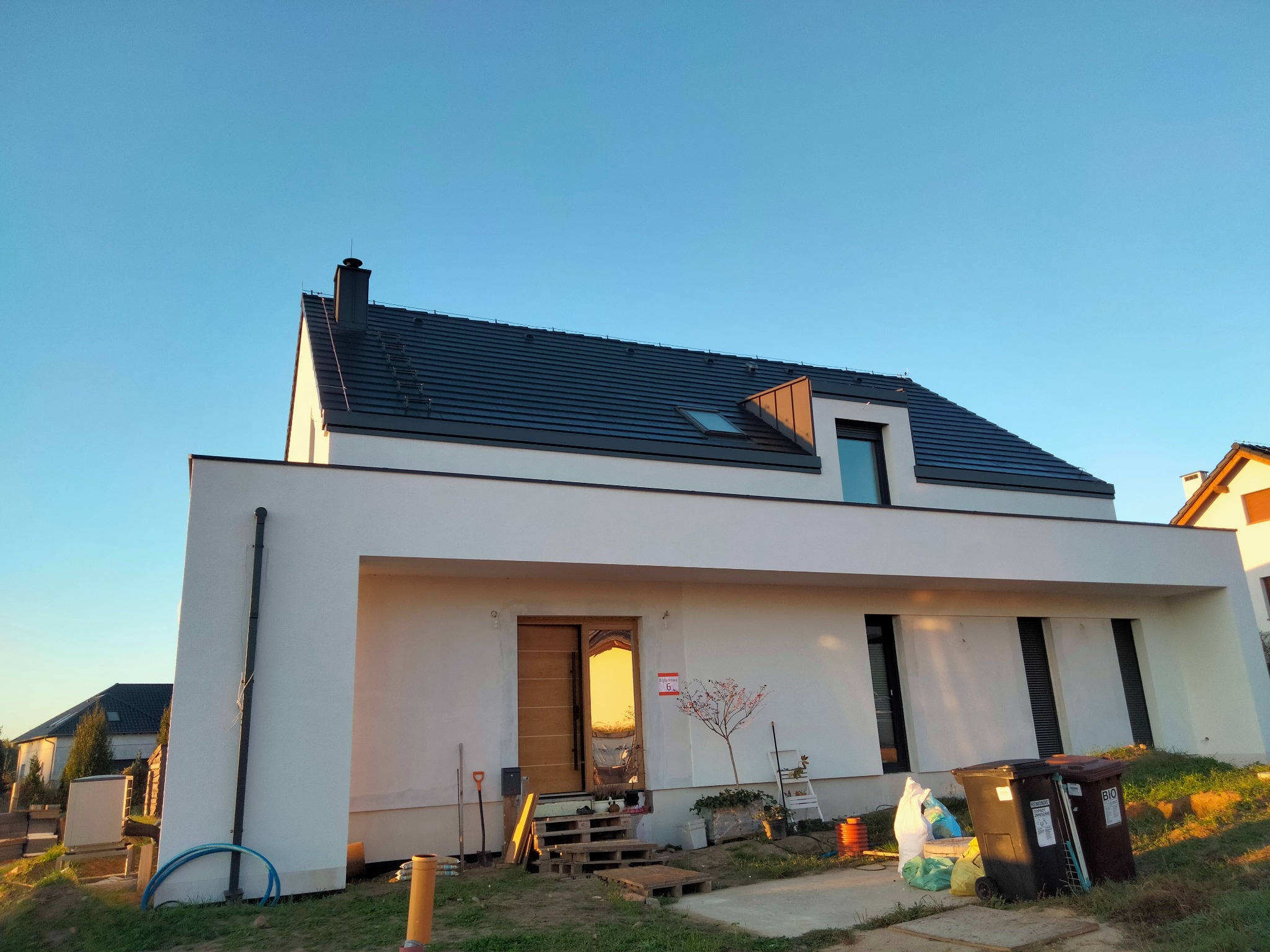 Nowoczesny dom jednorodzinny z ciemną dachówką i białymi ścianami w promieniach zachodzącego słońca. Widoczne detale wykończenia dachu i elewacji.