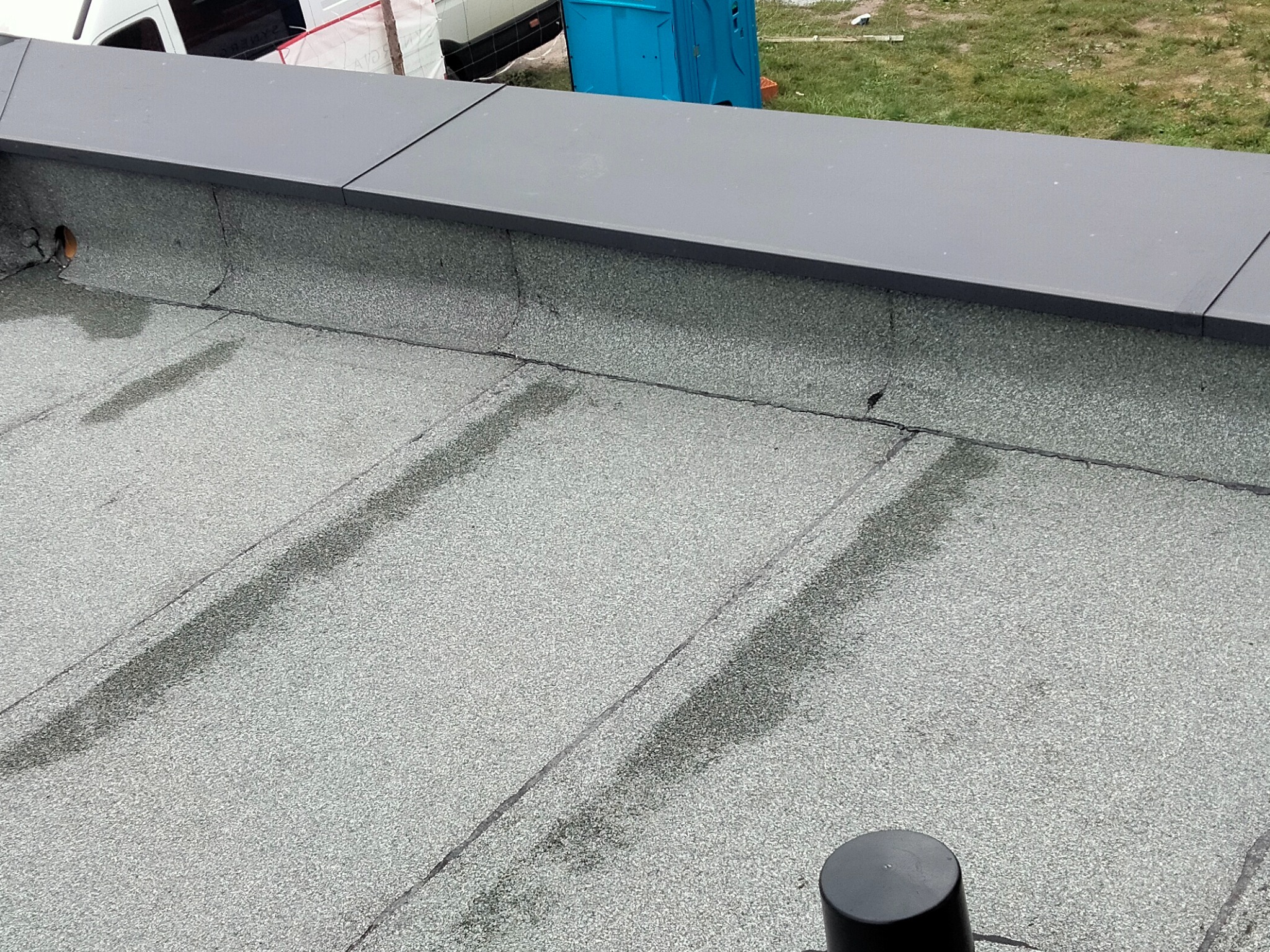 Szary, płaski dach pokryty papą z widocznymi śladami wilgoci i elementami obróbki blacharskiej. W tle fragment pojazdu i przenośna toaleta.
