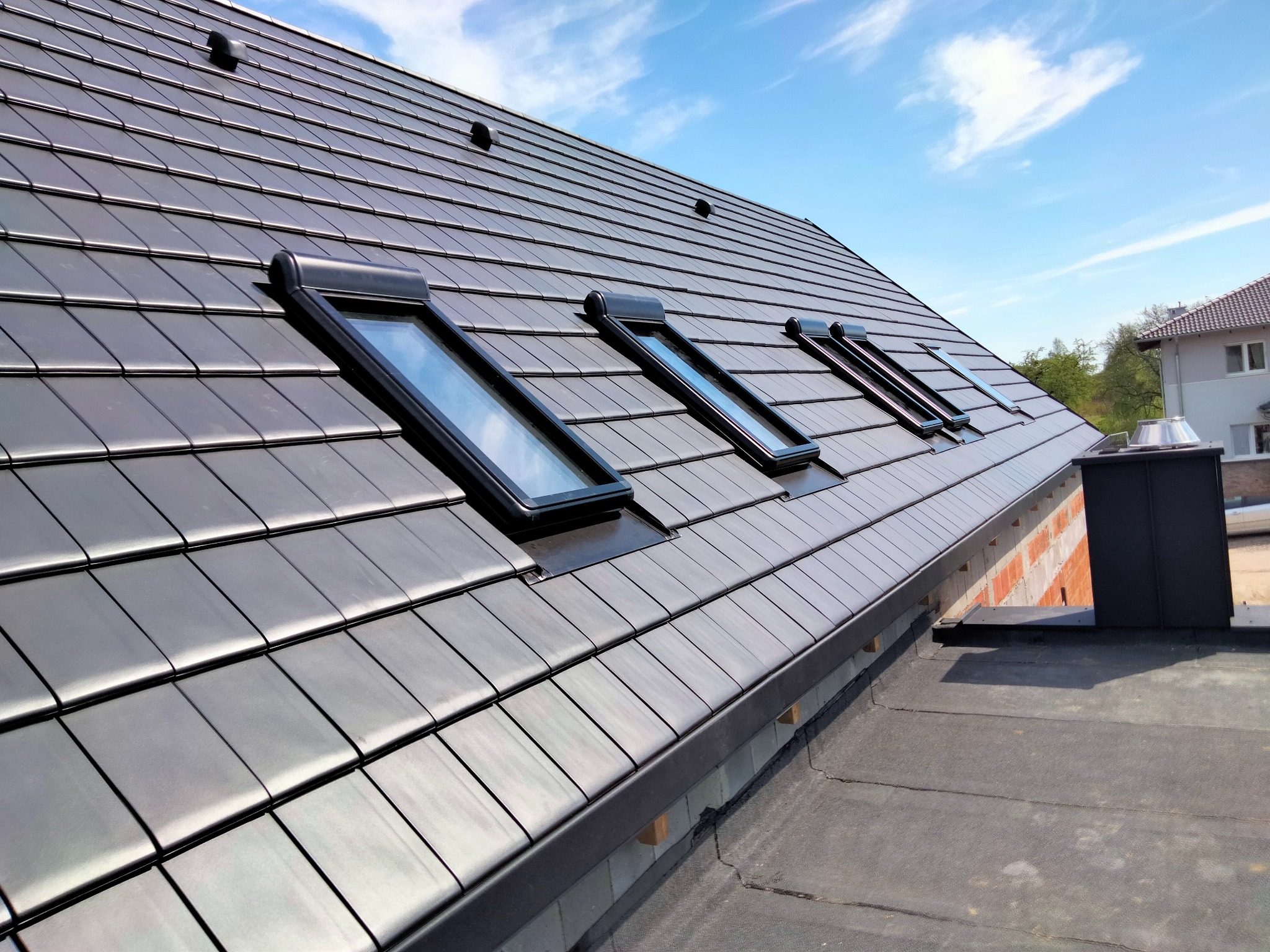 Czarny dach z dachówki ceramicznej z czterema oknami dachowymi, widok z dołu pod kątem. Na drugim planie błękitne niebo i fragment budynku.