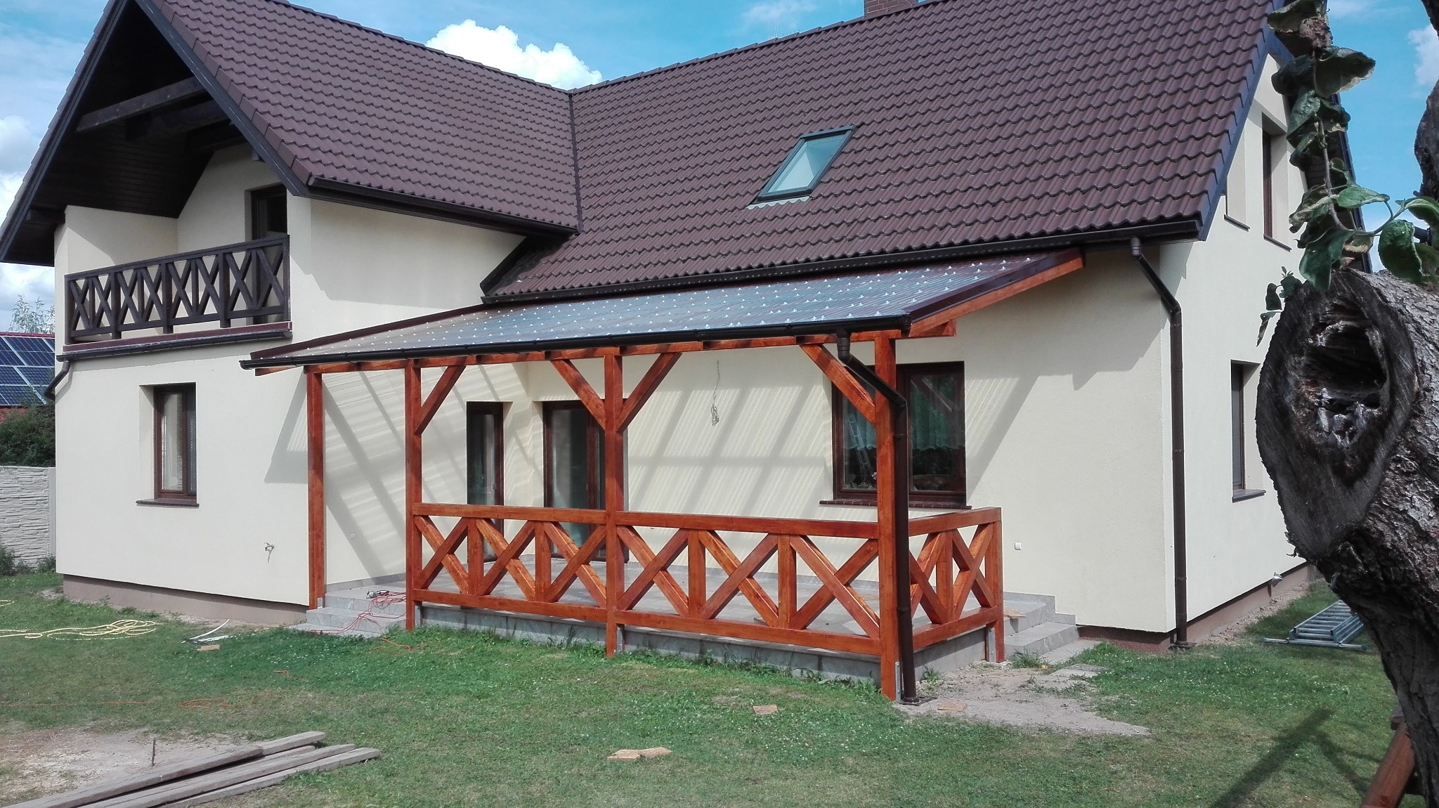 Dom z brązowym dachem i drewnianą pergolą z przezroczystym zadaszeniem. Widoczne rynny w kolorze dachu. Elementy drewniane pomalowane na brązowo.