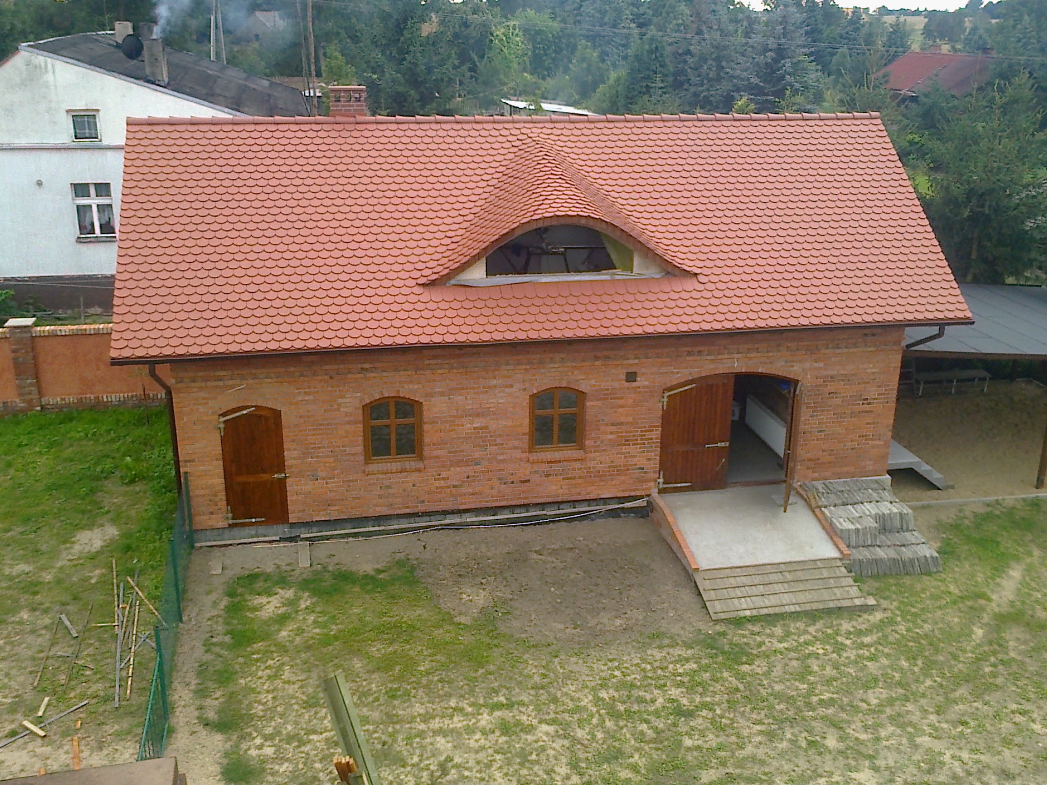 Ceglany budynek z nowym dachem z dachówki ceramicznej w odcieniach czerwieni, z oknem w kształcie łuku na poddaszu. Widok z góry, otoczenie zielone.