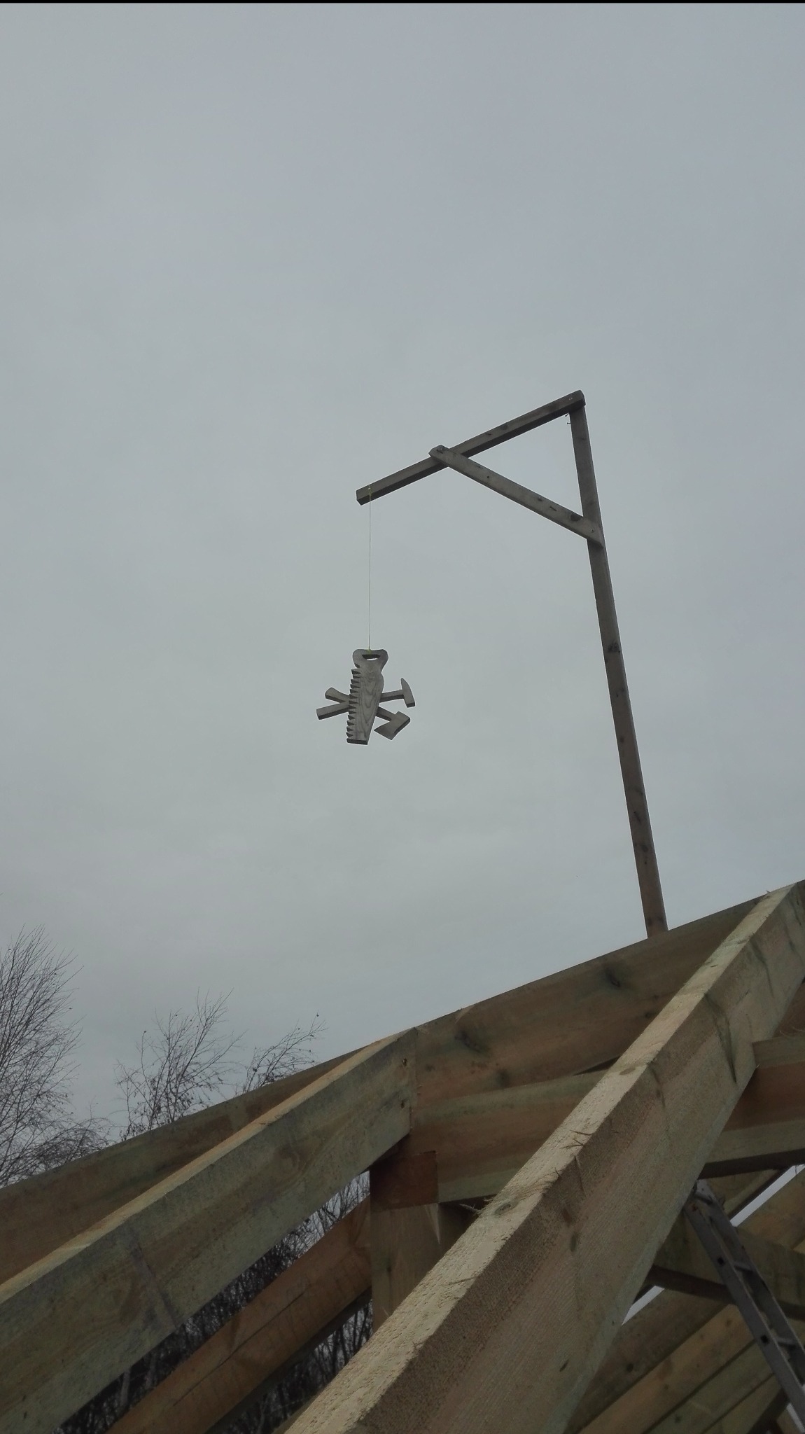 Wiązary dachowe z surowego drewna, w tle szare niebo. Na drewnianym wysięgniku zawieszona ozdoba z narzędzi dekarskich: piła, młotki, siekiera.