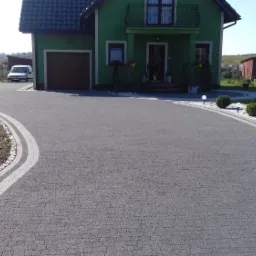 Szeroki podjazd z szarej kostki brukowej prowadzący do zielonego domu z garażem, obrzeża z jasnych kamieni i lamp solarnych, zadbany trawnik.