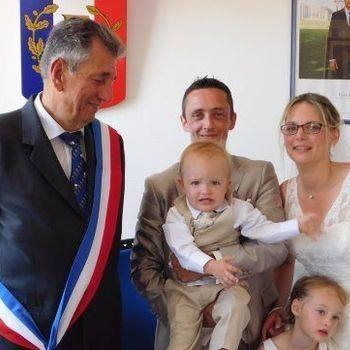 Uśmiechnięty mężczyzna w garniturze z szarfą w barwach flagi Francji stoi obok rodziny: mężczyzny trzymającego małe dziecko, kobiety w białej sukience i małej dziewczynki. W tle flaga i portret.