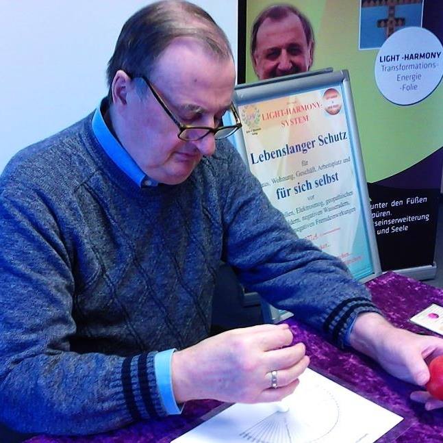 Mężczyzna w swetrze z geometrycznym wzorem, z obrączką na palcu, używa wahadełka nad diagramem na stole. W tle plakat z napisem 'Lebenslanger Schutz' i wizerunkiem innej osoby.