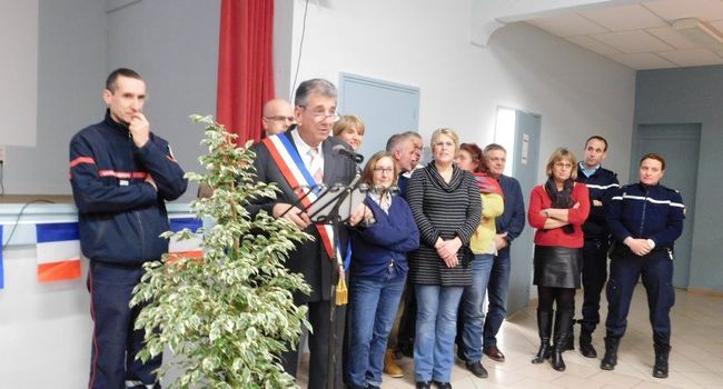 Burmistrz w szarfie przemawia do zgromadzonych, za nim rada miejska, obok delegacja żandarmerii i strażacy, na pierwszym planie roślina doniczkowa, w tle flaga Francji.