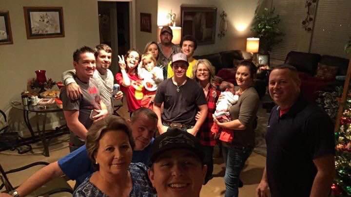 Duża grupa osób pozuje do selfie w salonie z choinką bożonarodzeniową w tle, część osób trzyma napoje i małe dzieci.