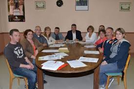 Grupa osób siedząca przy owalnym stole w pomieszczeniu, omawiająca dokumenty; na ścianie w tle wiszą dwa obrazy w ramach.