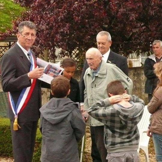 Uroczystość na zewnątrz: mężczyzna w garniturze z szarfą wręcza dokument starszemu mężczyźnie w otoczeniu dzieci i innych dorosłych.