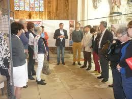 Grupa osób zgromadzona w przestronnym wnętrzu, prawdopodobnie podczas prezentacji lub wykładu. Mężczyzna w centrum, ubrany w marynarkę i jeansy, trzyma kartkę i przemawia do słuchaczy. W tle...