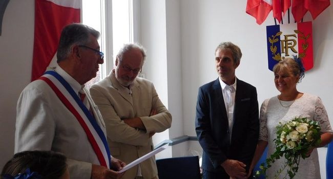 Urząd stanu cywilnego: urzędnik w szarfie odczytuje akt małżeństwa, obok świadkowie i para młoda z bukietem białych róż na tle francuskiej flagi i godła.