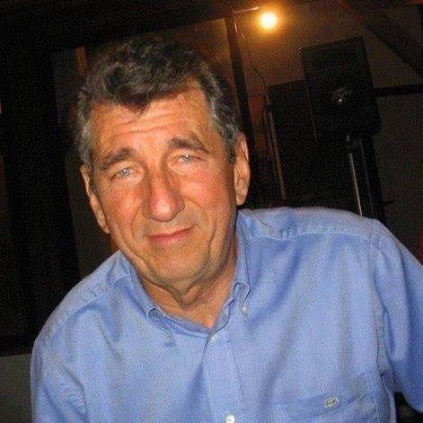 Portret starszego mężczyzny w niebieskiej koszuli na tle ciemnego wnętrza z widocznymi głośnikami i punktowym oświetleniem.