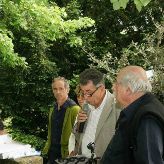 Prezentacja na zewnątrz: mężczyzna w okularach przemawia do grupy osób z mikrofonem na tle zieleni.