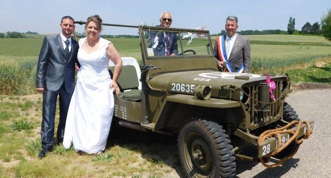 Para młoda pozuje obok wojskowego jeepa z numerem 20635, w tle zielone pola i drzewa, obok urzędnik stanu cywilnego z szarfą.