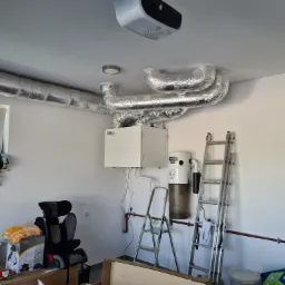 Instalacja rekuperacji w garażu: widoczne srebrne rury, centrala rekuperacyjna WanAs, drabiny i przedmioty codziennego użytku w tle.