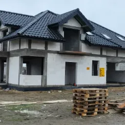 Dom w trakcie budowy z czarną dachówką karpiówką, widoczne okna dachowe, szary tynk na fundamentach, paleta i deski leżące na ziemi.