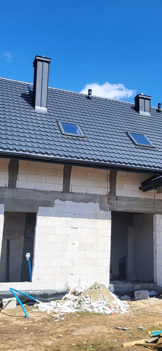 Dom w trakcie budowy z szarym dachem pokrytym dachówką, widoczne dwa okna dachowe i kominy, ściany z białego bloczka.