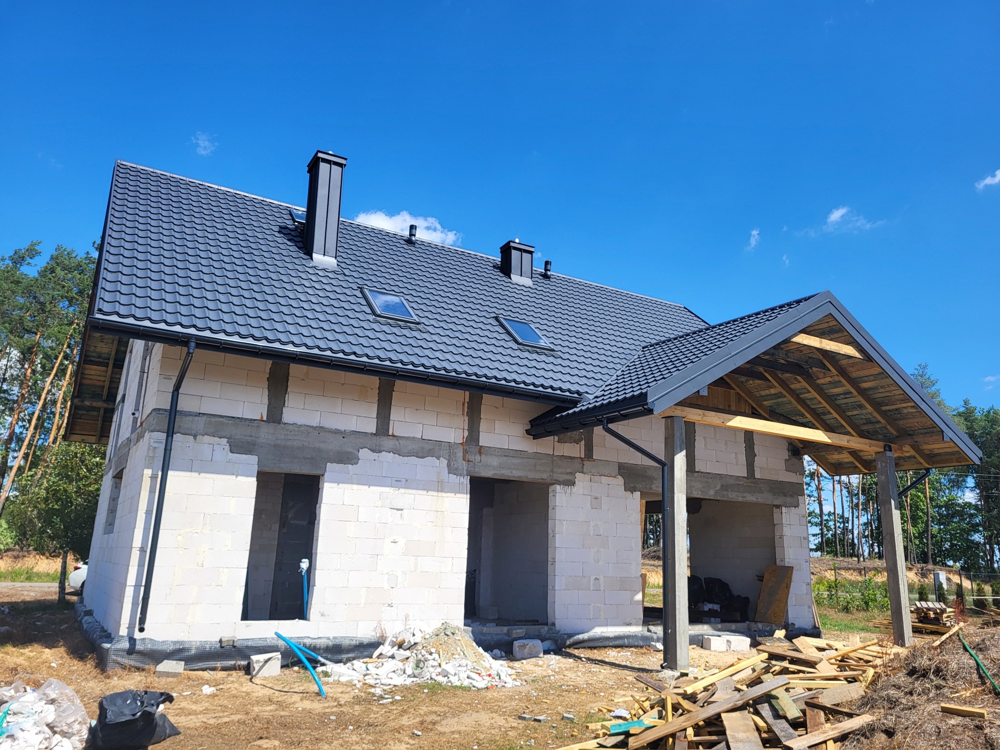 Dom w trakcie budowy z czarnym dachem z dachówki, widoczne kominy i okna dachowe, ściany z białego bloczka, zadaszenie tarasu w trakcie budowy, niebieskie niebo w tle.