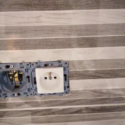 Gniazdka elektryczne podwójne, jedno z widocznymi przewodami, zamontowane na ścianie wyłożonej płytkami imitującymi drewno, w trakcie prac instalacyjnych w Tarnowskich Górach.