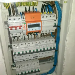 Rozdzielnica elektryczna z zainstalowanymi bezpiecznikami, licznikiem energii i okablowaniem, widoczne opisy bezpieczników na naklejkach.