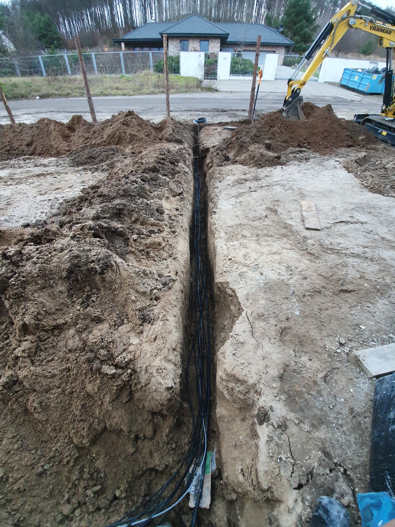 Wykop w ziemi z ułożonymi przewodami elektrycznymi, widoczny żółty minikoparka Yanmar i dom w tle.