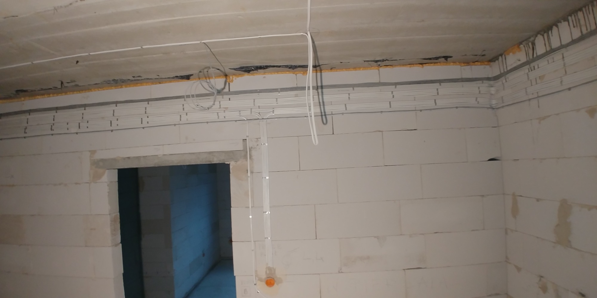 Wnętrze w stanie surowym z widoczną instalacją elektryczną: kable prowadzone w korytach na ścianach i suficie, widoczne puszki instalacyjne, ściany z białych bloczków.