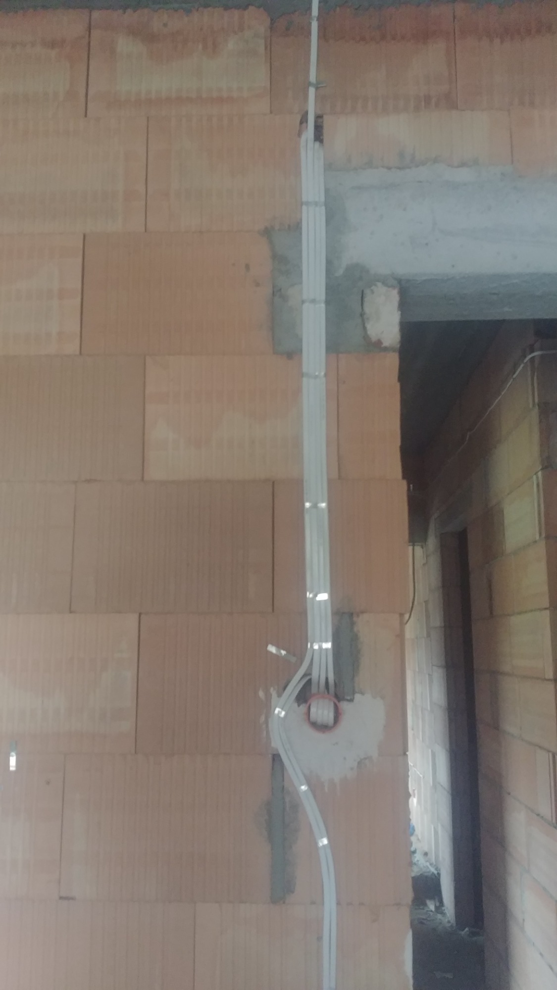 Ściana z cegły ceramicznej z pionowo ułożonymi przewodami elektrycznymi w białych peszlach, przymocowanymi metalowymi uchwytami, widoczna puszka instalacyjna.