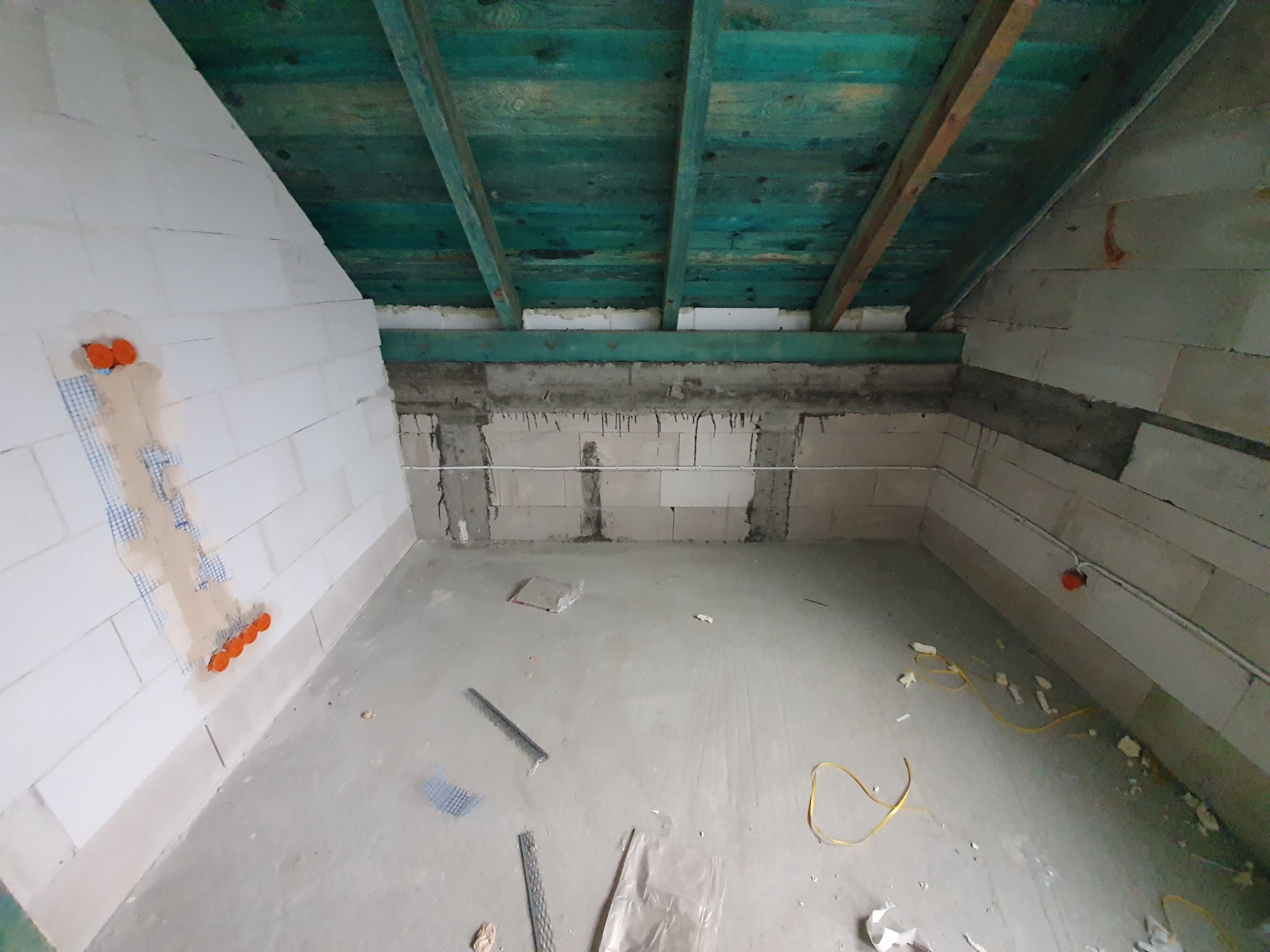 Surowy stan poddasza z widoczną instalacją elektryczną w ścianie z betonu komórkowego, zabezpieczoną siatką, oraz wystającymi pomarańczowymi puszkami. Drewniana, pomalowana na zielono konstrukcja...