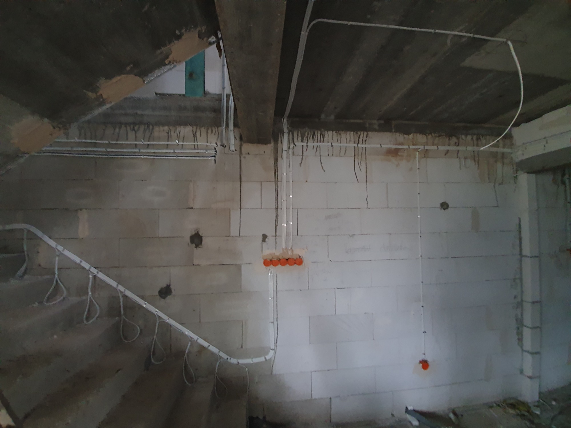 Surowy stan budynku w trakcie prac elektrycznych: widoczne przewody w peszlach na ścianach z betonu komórkowego, puszki instalacyjne w kolorze pomarańczowym, instalacja pod schodami.
