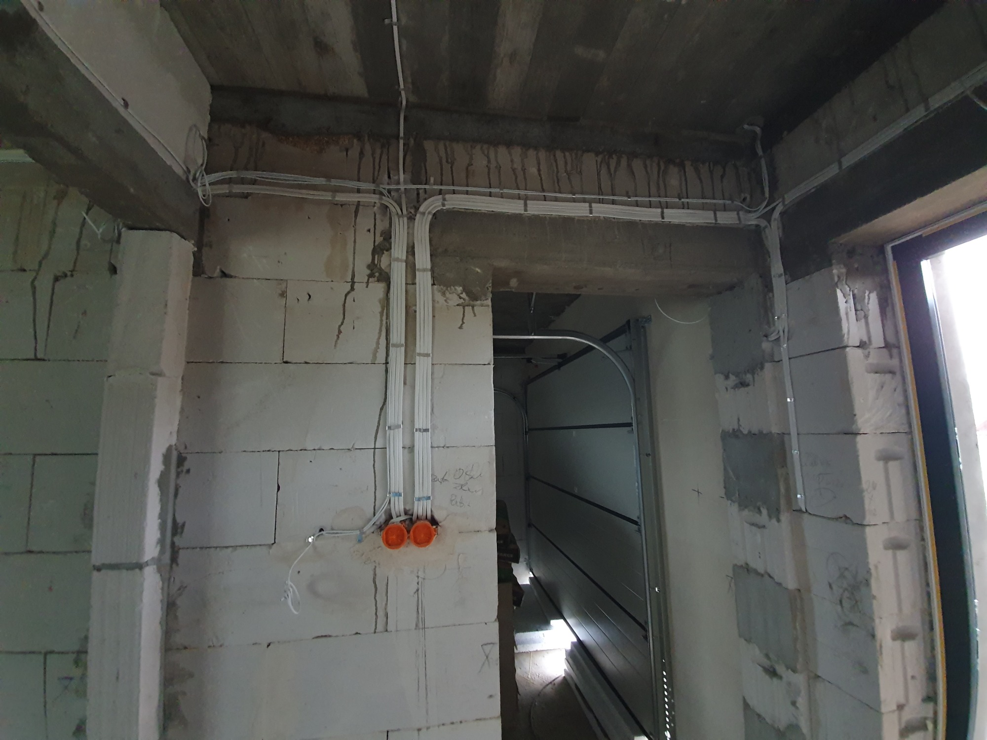 Surowy stan instalacji elektrycznej w nowym budynku: białe przewody prowadzone po ścianach z betonu komórkowego, dwie pomarańczowe puszki instalacyjne widoczne na ścianie, sufit betonowy...