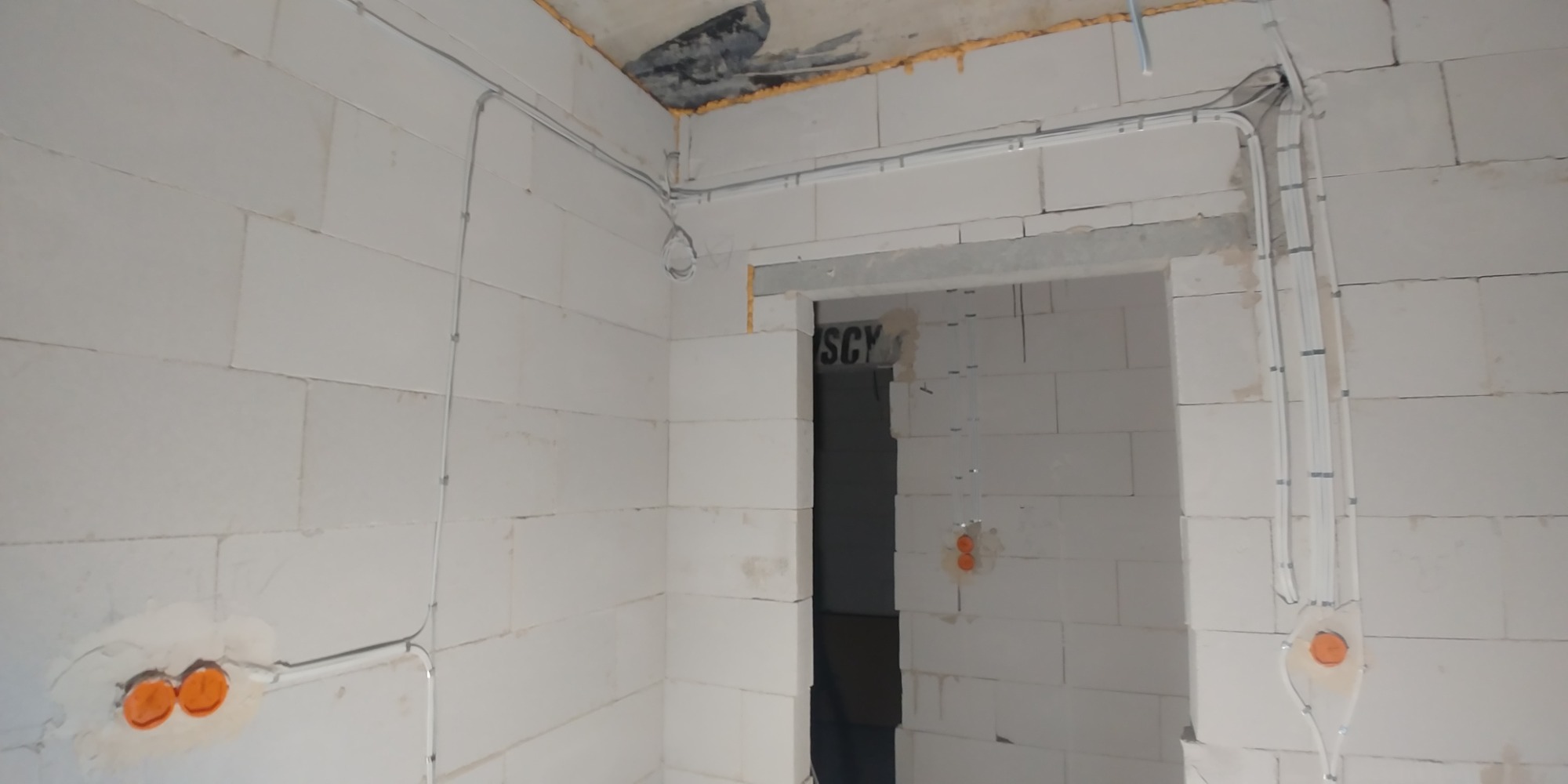 Surowy stan: Instalacja elektryczna w budynku z białego bloczka, widoczne kable prowadzone natynkowo, puszki podtynkowe z pomarańczowymi osłonami, niezabezpieczone otwory w suficie.