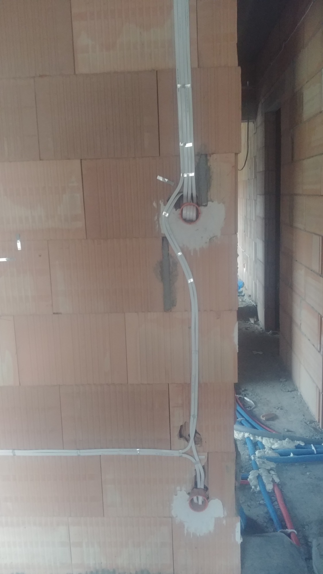 Wewnętrzne instalacje elektryczne w trakcie montażu w nowym budynku z czerwonej cegły, widoczne kable i puszki elektryczne w ścianie, obok instalacje wodne.