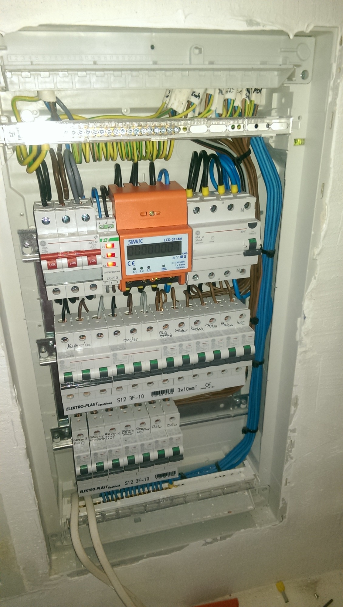 Rozdzielnica elektryczna z zainstalowanymi bezpiecznikami, licznikiem energii i okablowaniem, widoczne opisy bezpieczników na naklejkach.