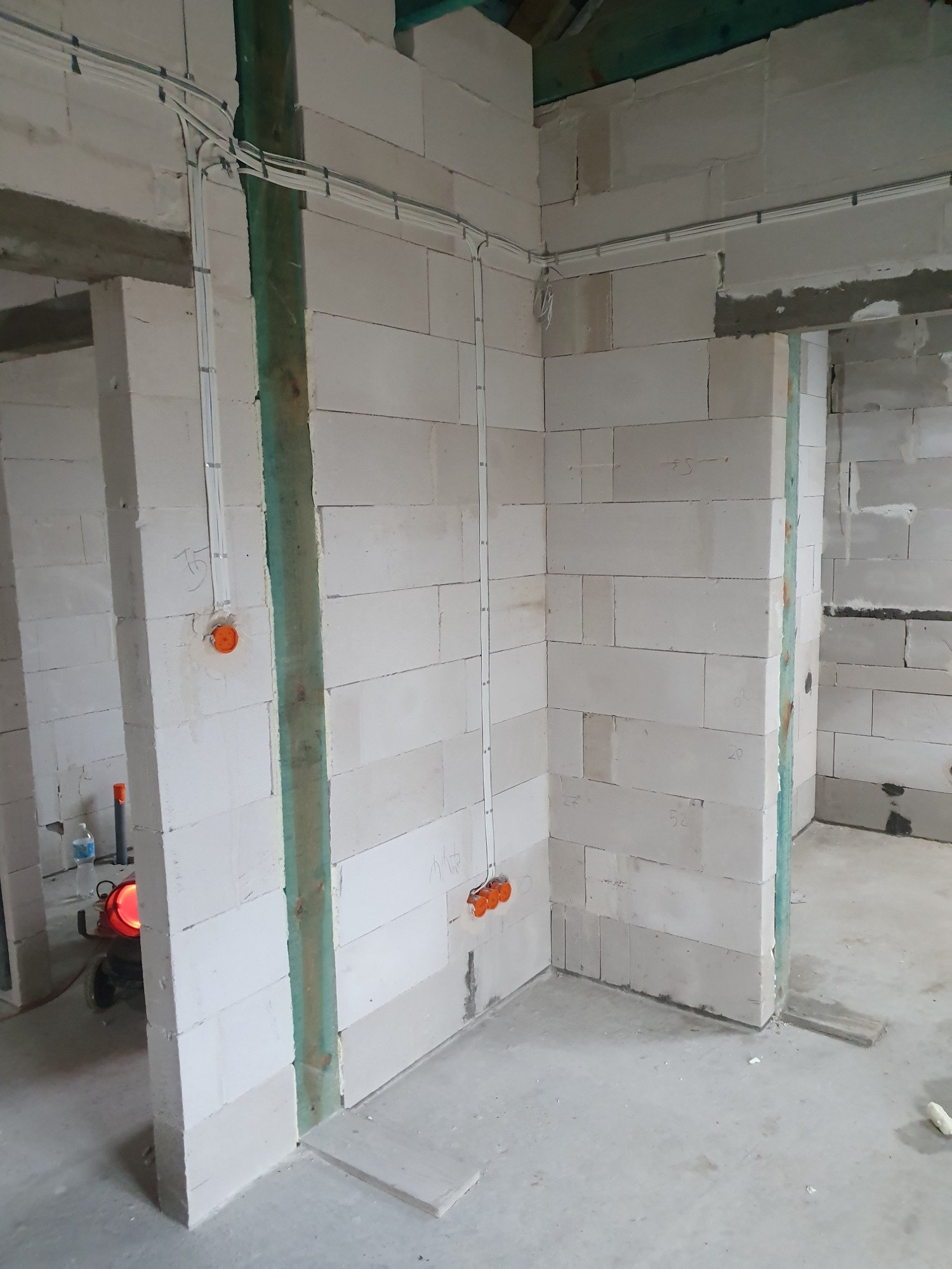 Wnętrze w trakcie budowy z białych bloczków, widoczne kable elektryczne prowadzone po ścianach i puszki instalacyjne w kolorze pomarańczowym, zielone drewniane elementy konstrukcyjne, surowy beton...