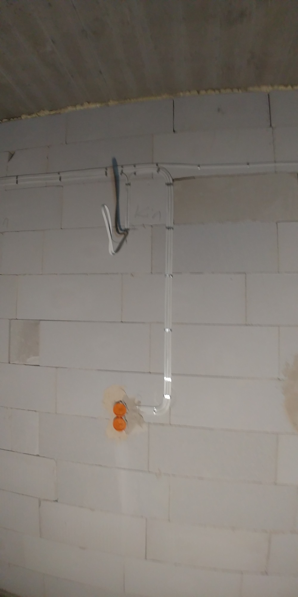 Szara ściana z betonu komórkowego z zamontowanymi białymi rurkami instalacji elektrycznej, widoczne dwa pomarańczowe gniazda elektryczne w ścianie.