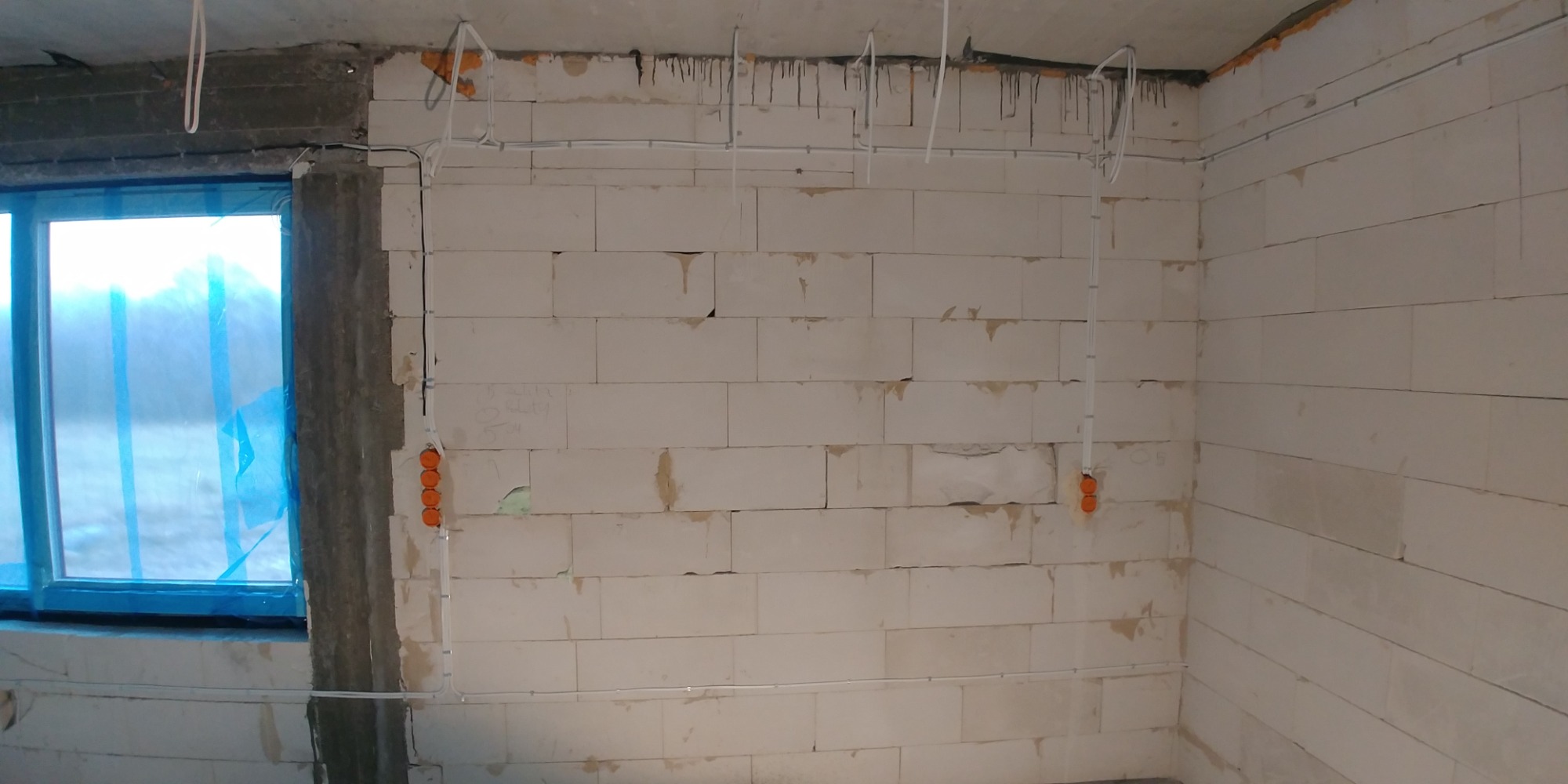 Surowy stan: Ściany z betonu komórkowego w trakcie rozprowadzania instalacji elektrycznej, widoczne kable i pomarańczowe puszki podtynkowe, okno zabezpieczone folią.