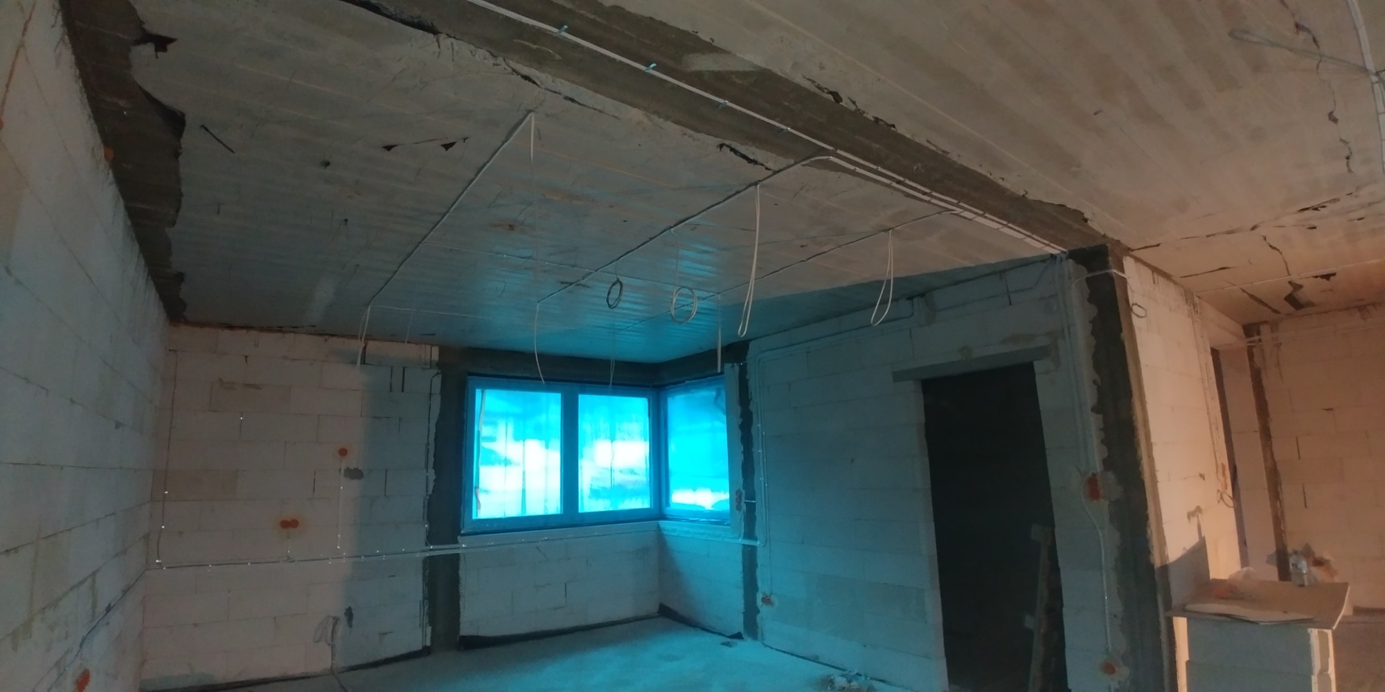 Surowy stan wnętrza z widoczną instalacją elektryczną: przewody prowadzone po suficie i ścianach, puszki elektryczne w murze z białych bloczków, okno zabezpieczone folią.