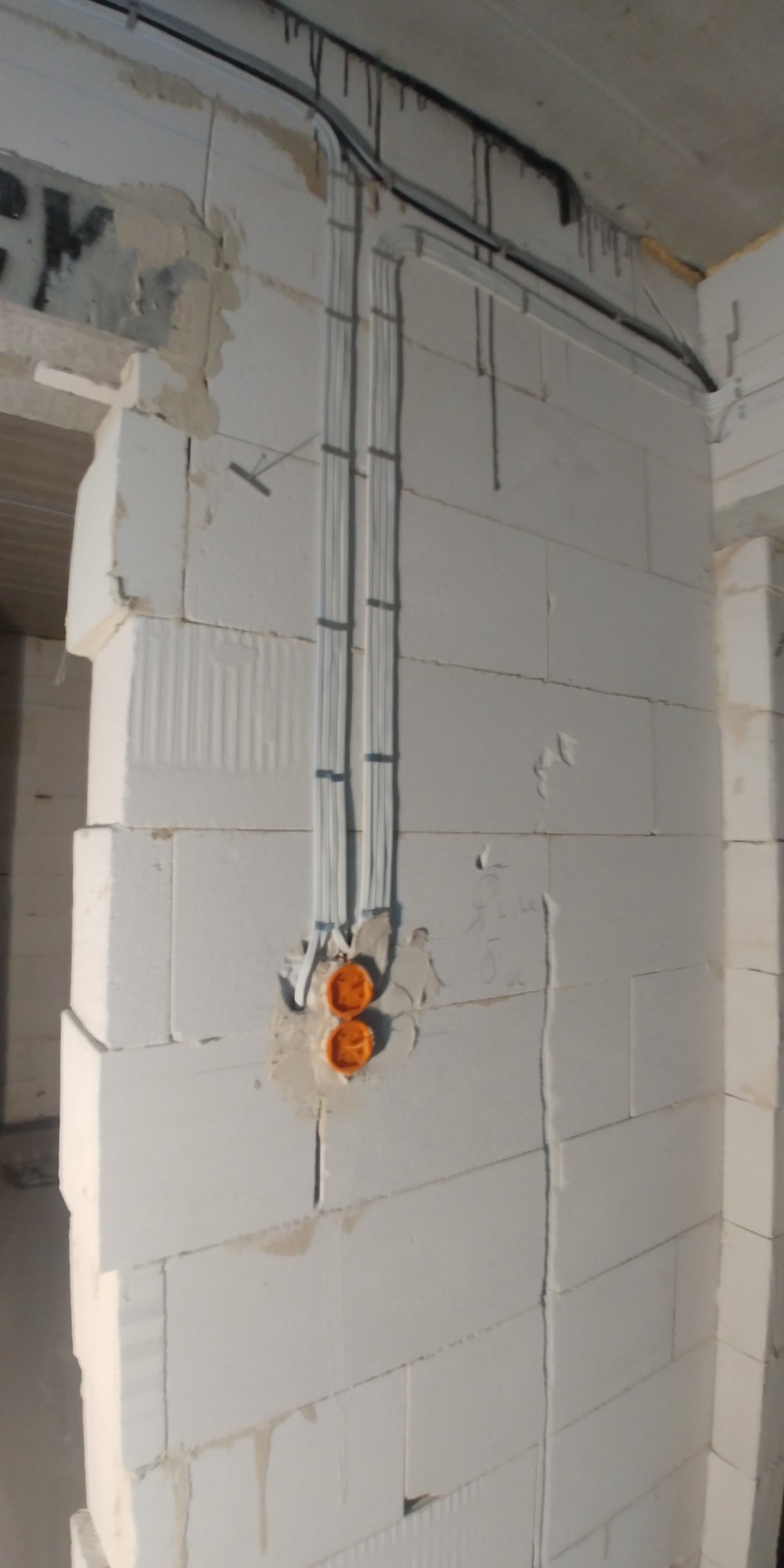 Instalacja elektryczna w surowym stanie: kable w białych peszlach przymocowane do ściany z białych bloczków, puszki elektryczne w kolorze pomarańczowym, widoczne ślady po montażu.