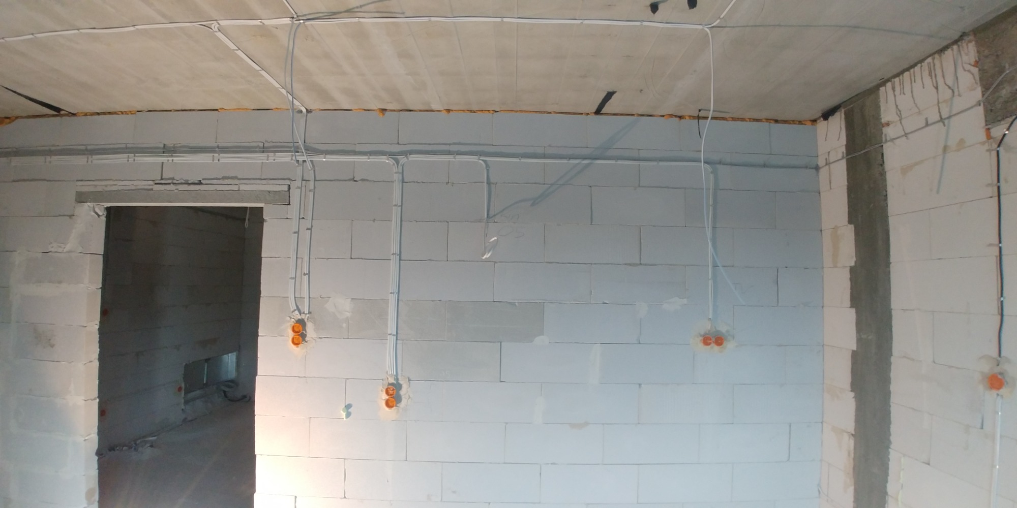 Surowy stan wnętrza z białych bloczków, widoczne kable elektryczne prowadzone po ścianach i suficie, pomarańczowe puszki pod gniazdka, widok na otwór drzwiowy.