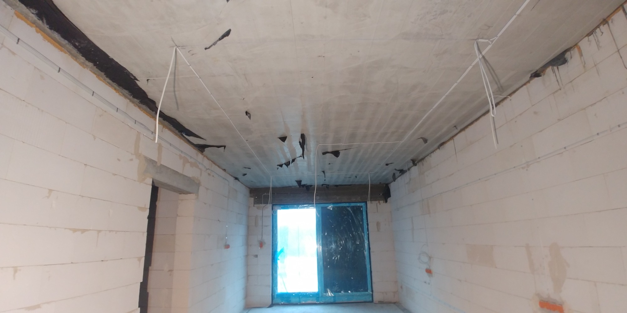 Wnętrze w stanie surowym z widocznymi przewodami elektrycznymi pod sufitem, przygotowane do dalszych prac wykończeniowych.