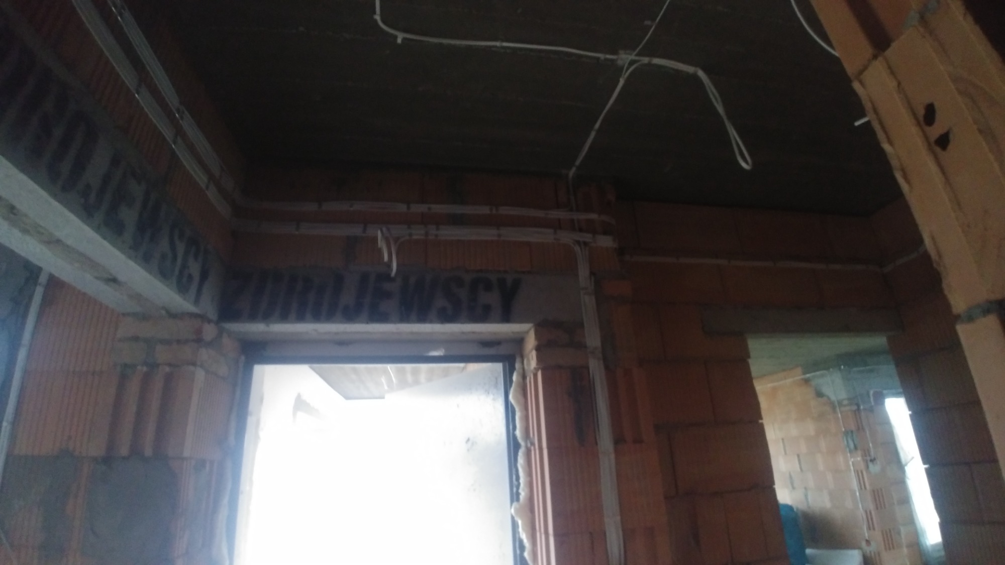 Wnętrze w stanie surowym: widoczne przewody elektryczne poprowadzone po suficie i ścianach, ściany z czerwonej cegły, otwory drzwiowe bez ościeżnic.