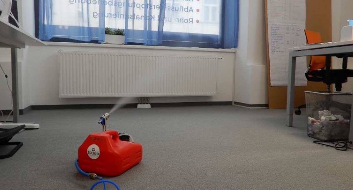 Czerwone urządzenie zamgławiające marki Soluto rozpyla środek dezynfekujący w biurze, widoczny kaloryfer i okno z niebieskimi zasłonami w tle.