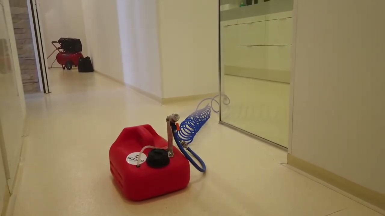 Czerwony pojemnik z rozpylaczem i niebieskim wężem na podłodze w sterylnym, białym pomieszczeniu, w tle kompresor powietrza, prawdopodobnie podczas dezynfekcji metodą zamgławiania.