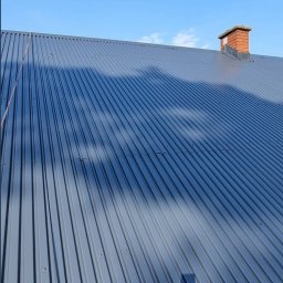 M-Color - Szary dach z blachy trapezowej w słoneczny dzień, widoczny ceglany komin. Czyste niebo w tle. Widoczne cienie na dachu.