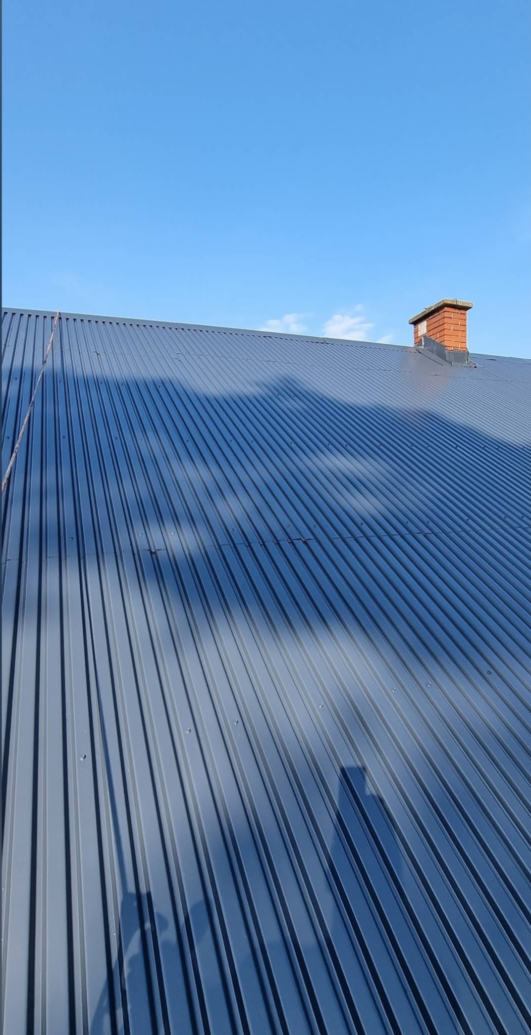 Szary dach z blachy trapezowej w słoneczny dzień, widoczny ceglany komin. Czyste niebo w tle. Widoczne cienie na dachu.