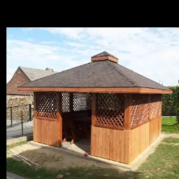 Drewniana altana z brązowym dachem z gontów bitumicznych, otoczona zielonym trawnikiem, widok z boku na tle błękitnego nieba z lekkimi chmurami.