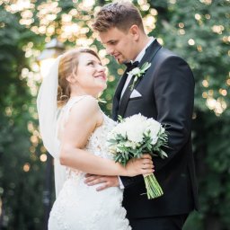 Natalia Nakonechna Photography - Uśmiechnięta panna młoda w koronkowej sukni spogląda na pana młodego w czarnym smokingu, trzymając bukiet białych kwiatów na tle rozmytych świateł.