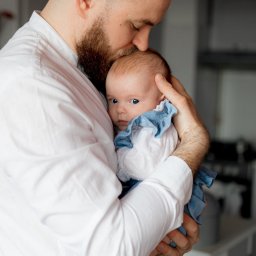Natalia Nakonechna Photography - Tata w białej koszuli delikatnie całuje w główkę niemowlę ubrane w błękitną sukienkę z falbankami, patrzące wprost w obiektyw