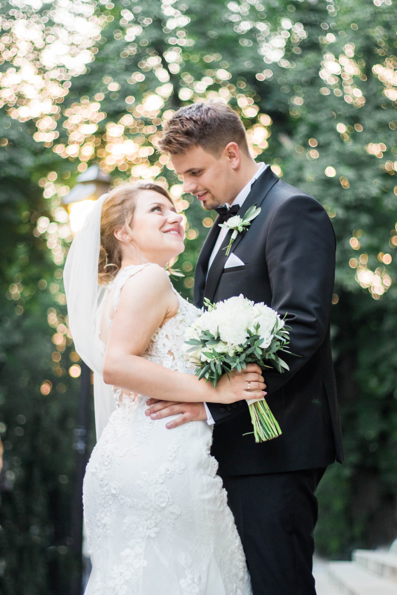 Uśmiechnięta panna młoda w koronkowej sukni spogląda na pana młodego w czarnym smokingu, trzymając bukiet białych kwiatów na tle rozmytych świateł.