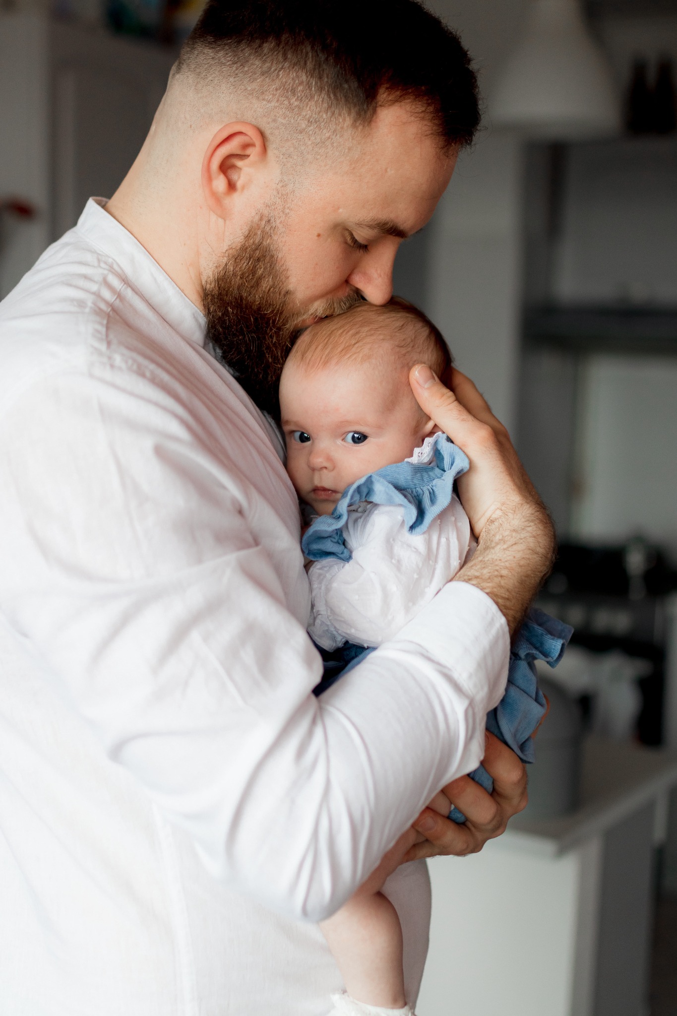 Tata w białej koszuli delikatnie całuje w główkę niemowlę ubrane w błękitną sukienkę z falbankami, patrzące wprost w obiektyw